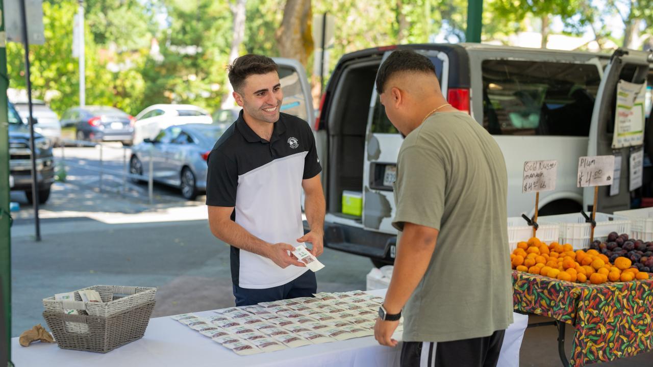 Dion Skaria, a food science major, sells his Fit Candy product at the Davis farmer's market. Photo by: Jael Mackendorf, UC Davis. A seller and a prospective buyer meet over a table at the Davis Farmer's market. Product is on the table.