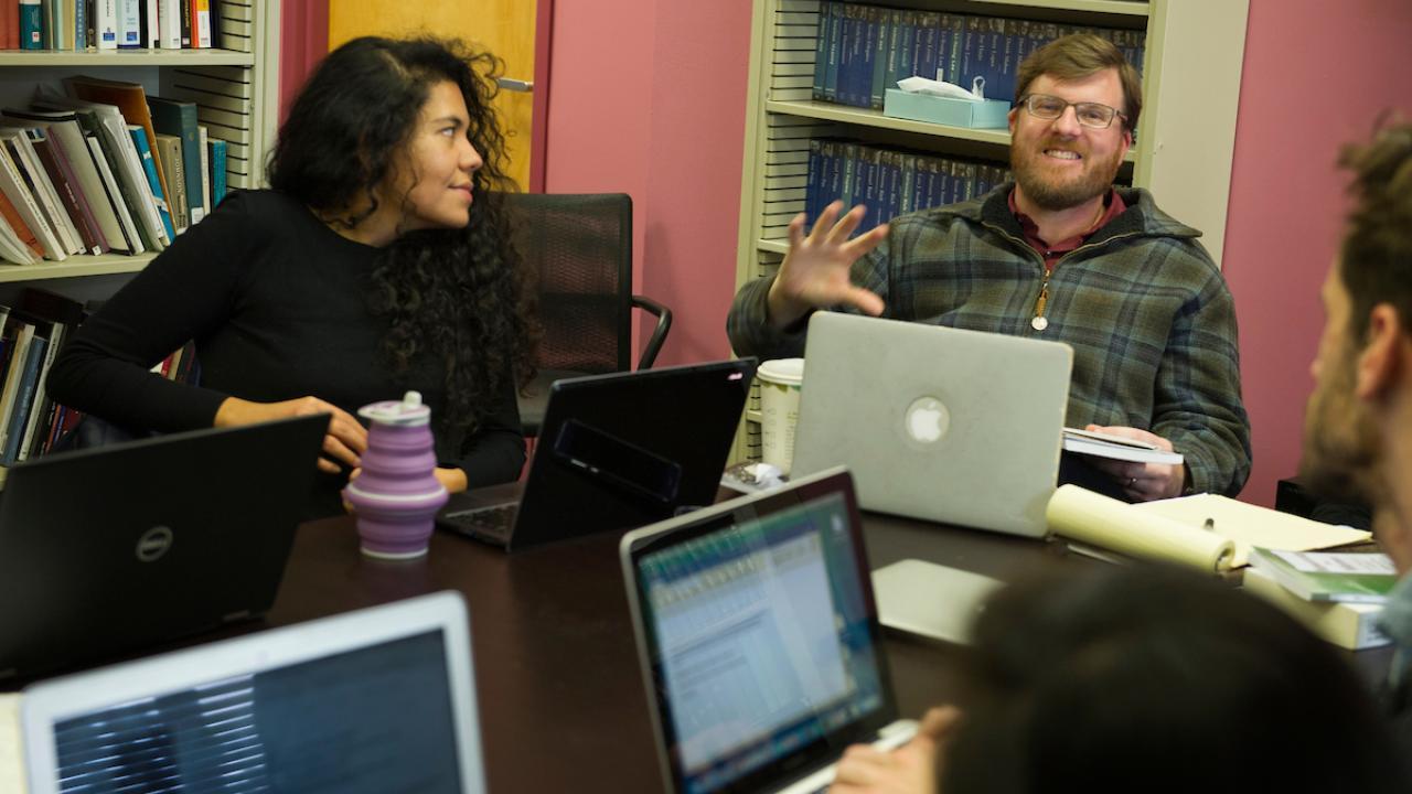 Three people sit at a table with laptops, meeting and talking.