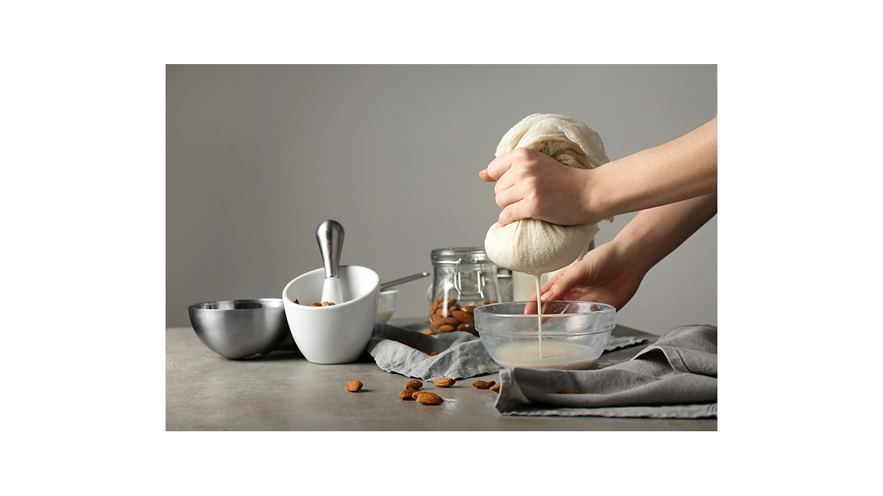 Hands squeeze almonds through cheesecloth into a clear bowl to make almond milk.